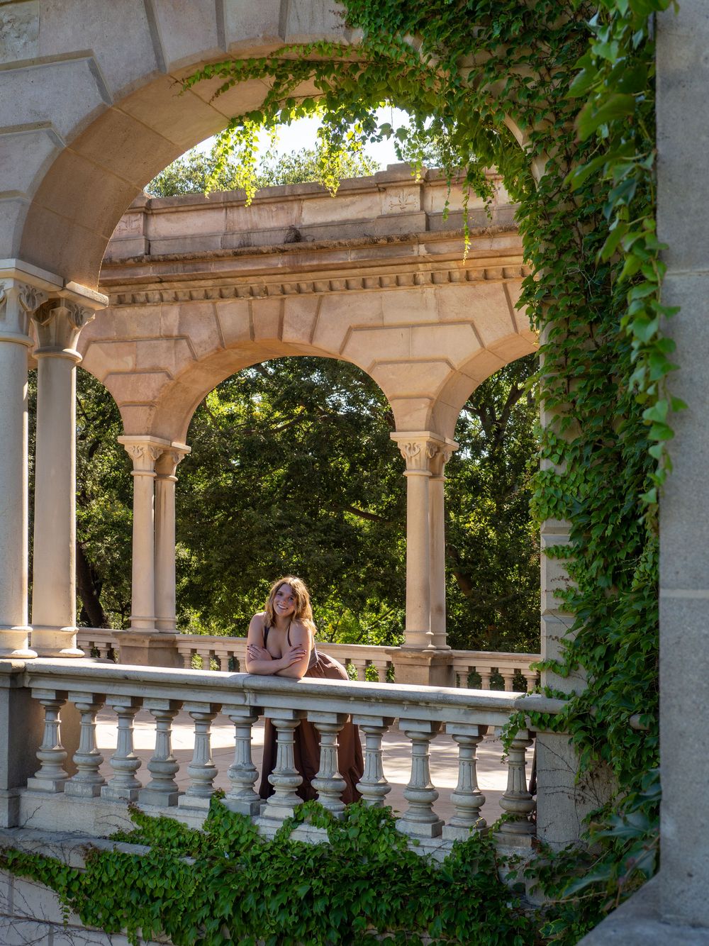Woman posing under ornate stone arches draped in ivy, with lush green trees visible through the classical colonnade.