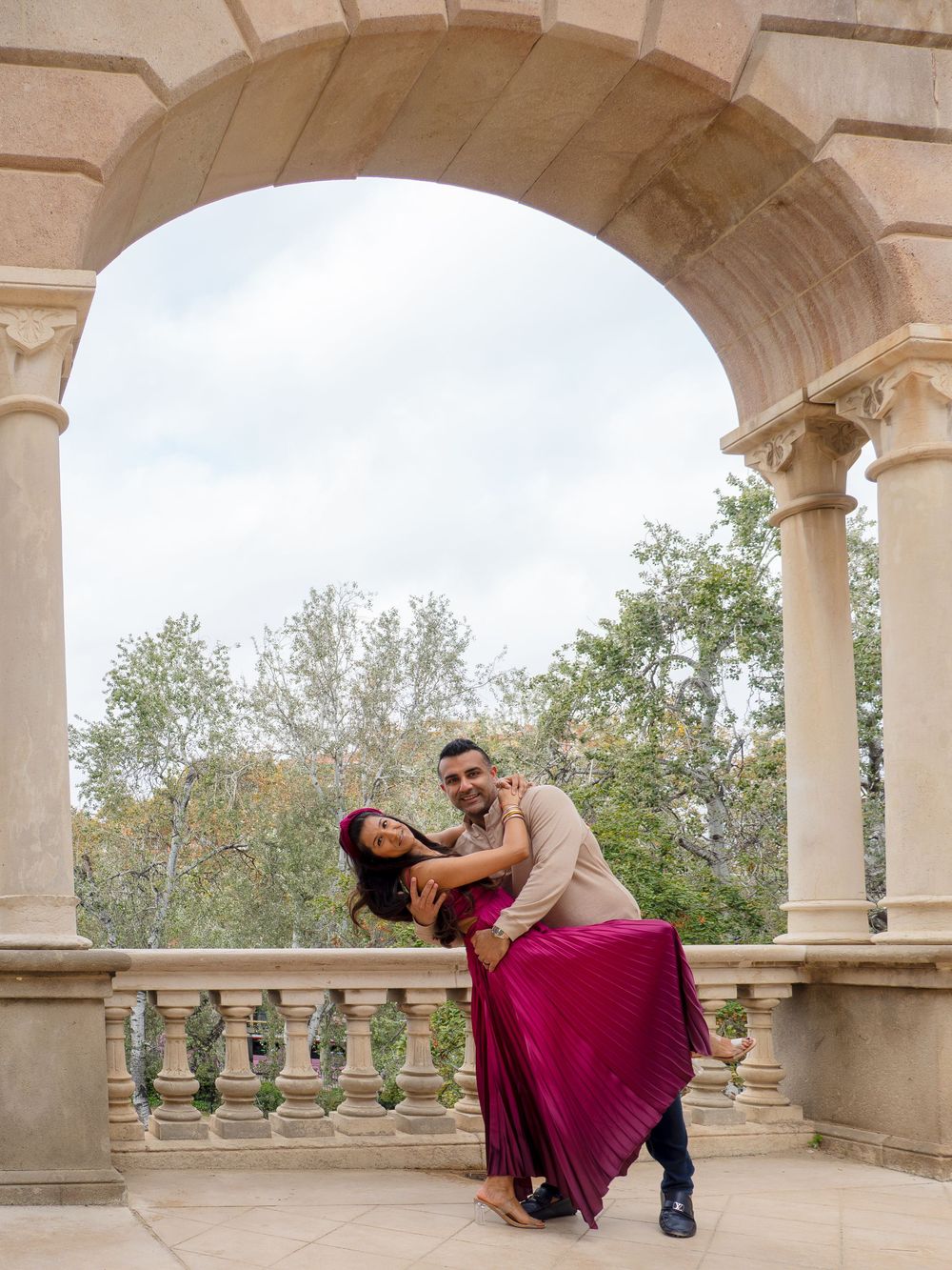 Couple dancing under stone arch on elegant balcony, woman in magenta gown, lush trees in background.