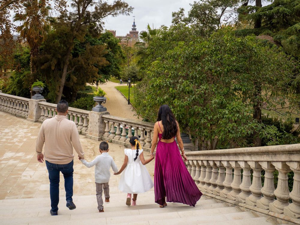 Family of four walking hand-in-hand along a scenic stone bridge path surrounded by lush green trees.