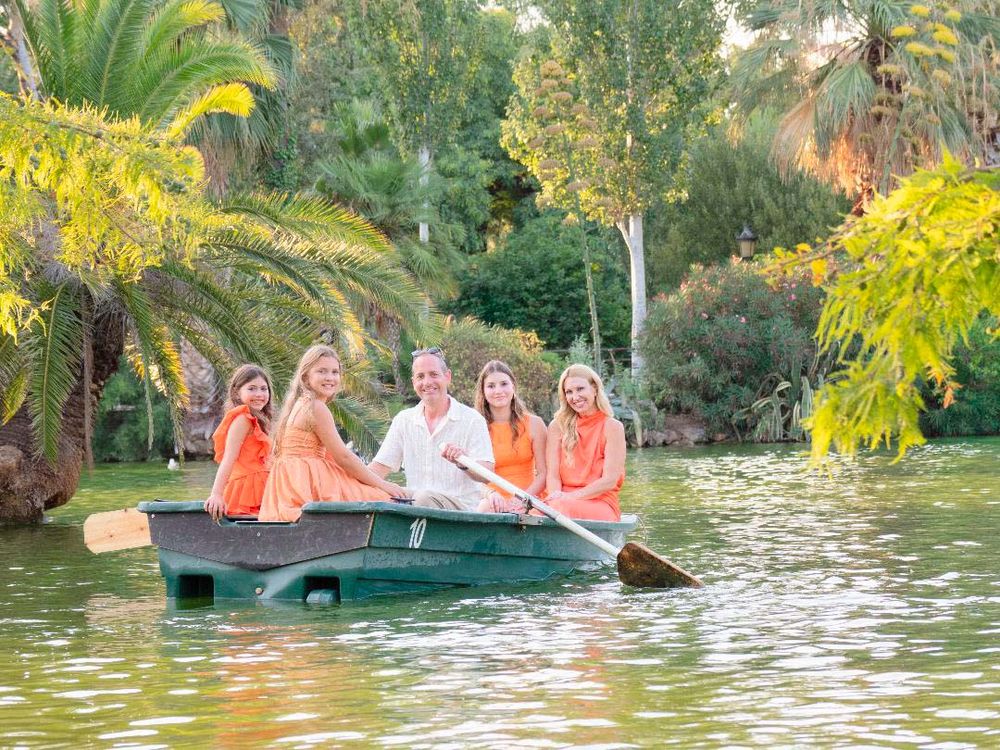 Family of five in matching orange outfits rowing a boat on a serene lake surrounded by lush green trees.