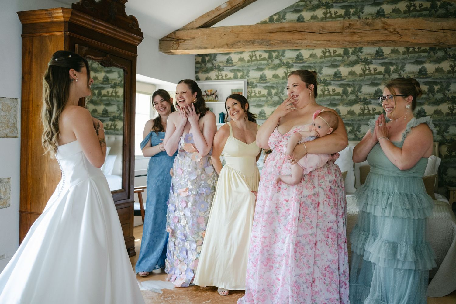 Bride in white gown shares a warm group hug with bridesmaids in a charming room with green toile wallpaper.