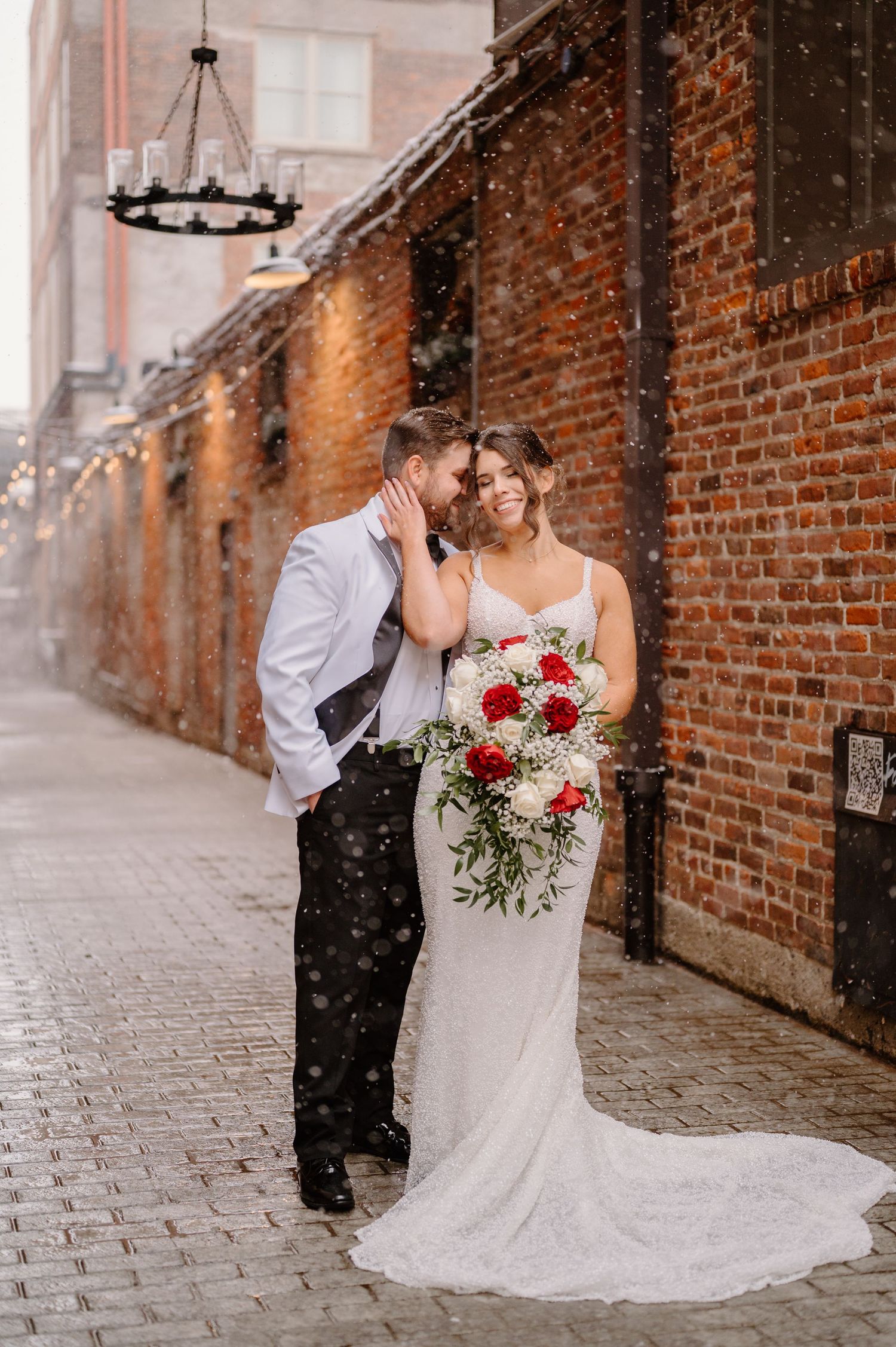 Bride and groom embrace in snowy alley beside brick wall, bride holding red and white bouquet in white lace gown.
