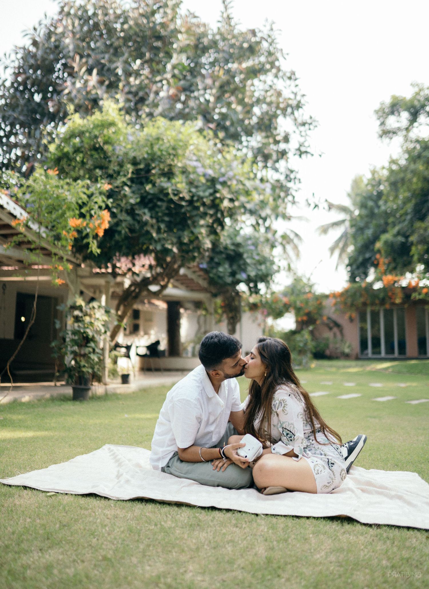 Couple sharing a romantic kiss while sitting on a blanket in a lush garden with trees and a house in the background.
