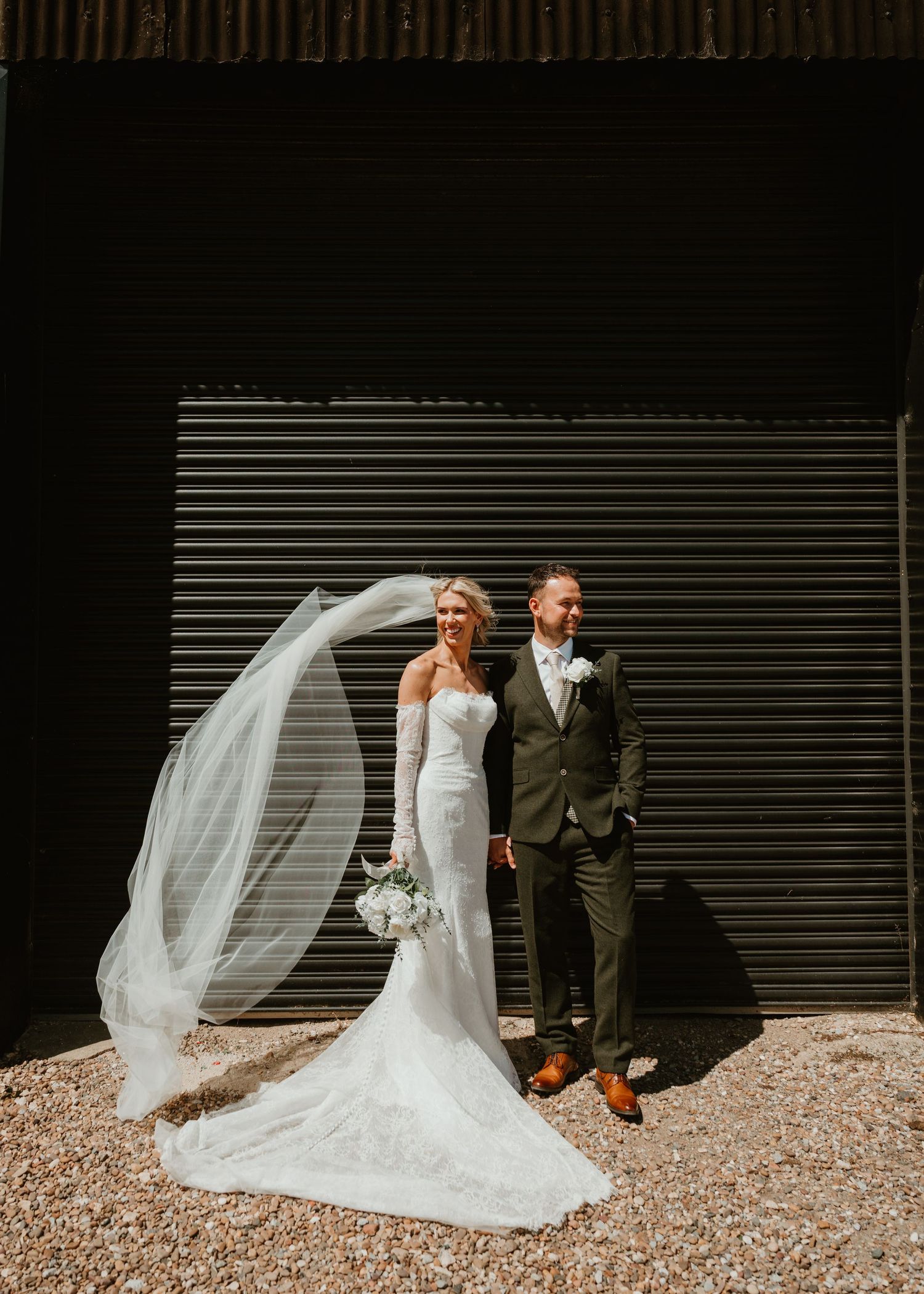 Bride in lace gown with flowing veil beside groom in olive suit against dark barn backdrop.