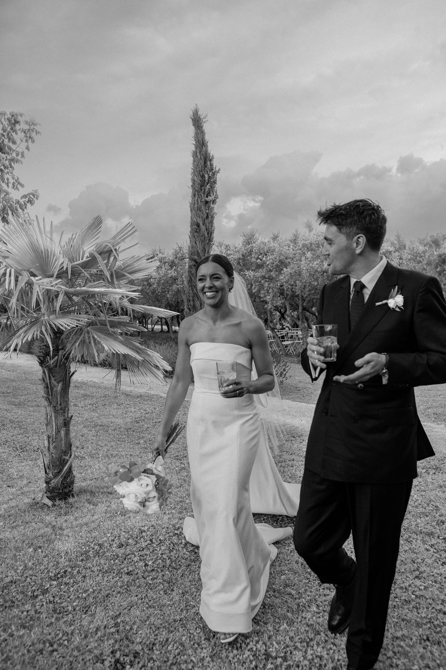 Black and white photo of a smiling bride in strapless gown and groom in suit walking outdoors at wedding. Bridal dress from Victor and Rolf