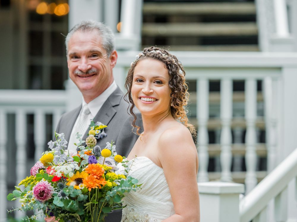 Smiling bride holding colorful wedding bouquet stands with groom on white porch steps.