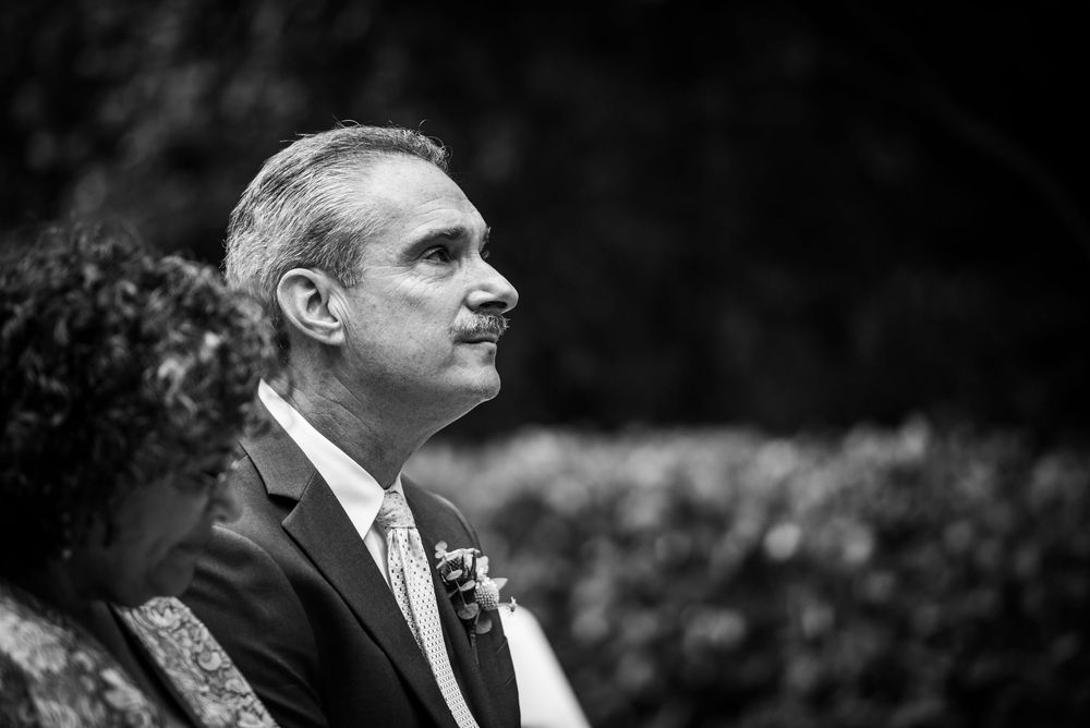 Elegant groom in suit with boutonniere gazing forward at outdoor wedding ceremony, black and white.