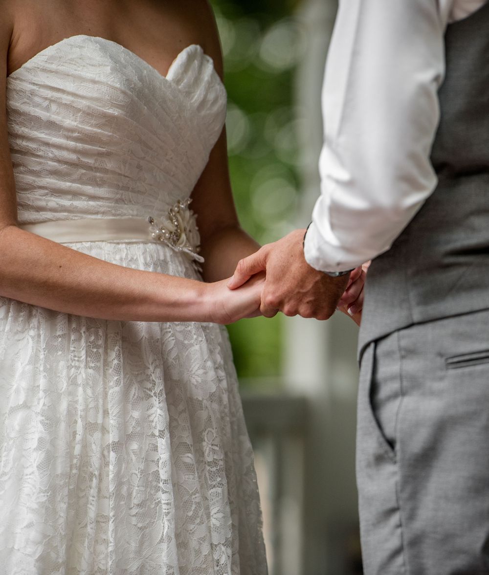 Bride and groom holding hands during wedding ceremony, bride in strapless lace gown, groom in gray suit.