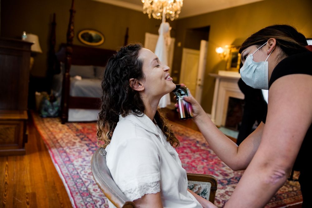 Bride getting makeup applied by masked artist in elegant bedroom before wedding ceremony.