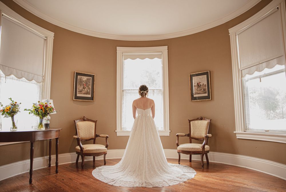 Elegant bride in white gown with long train standing in curved bridal suite facing windows.