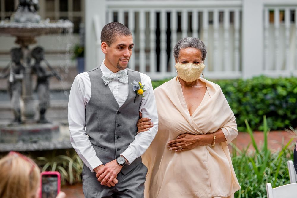 Groom in gray vest walks with masked woman in beige at outdoor wedding ceremony.
