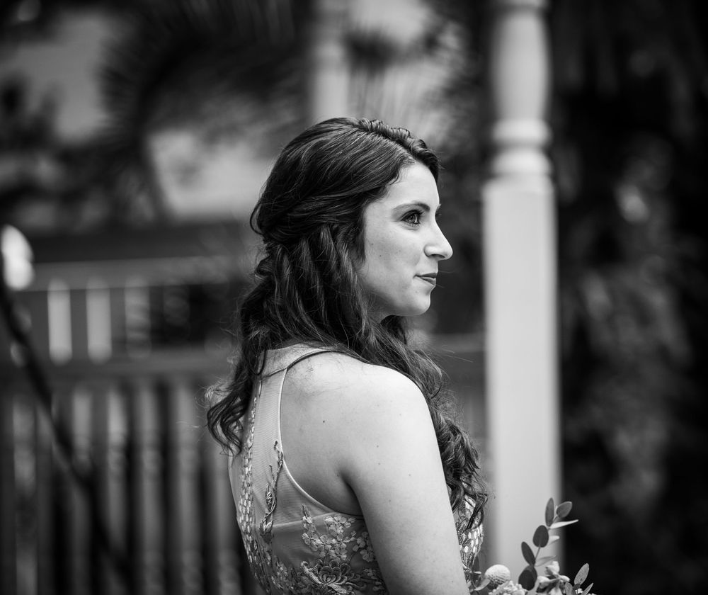 Black and white photo of a young woman in a formal gown with long wavy hair, posing outdoors at an elegant venue.