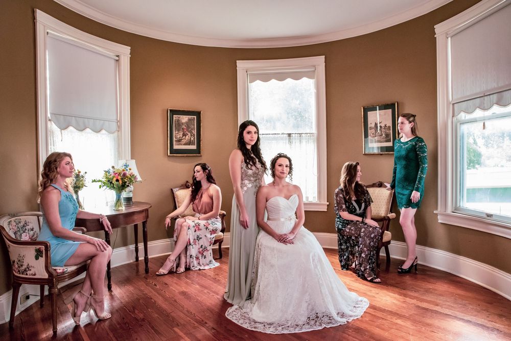 Bride in white gown surrounded by bridesmaids in elegant dresses in a warm-toned bay window room.
