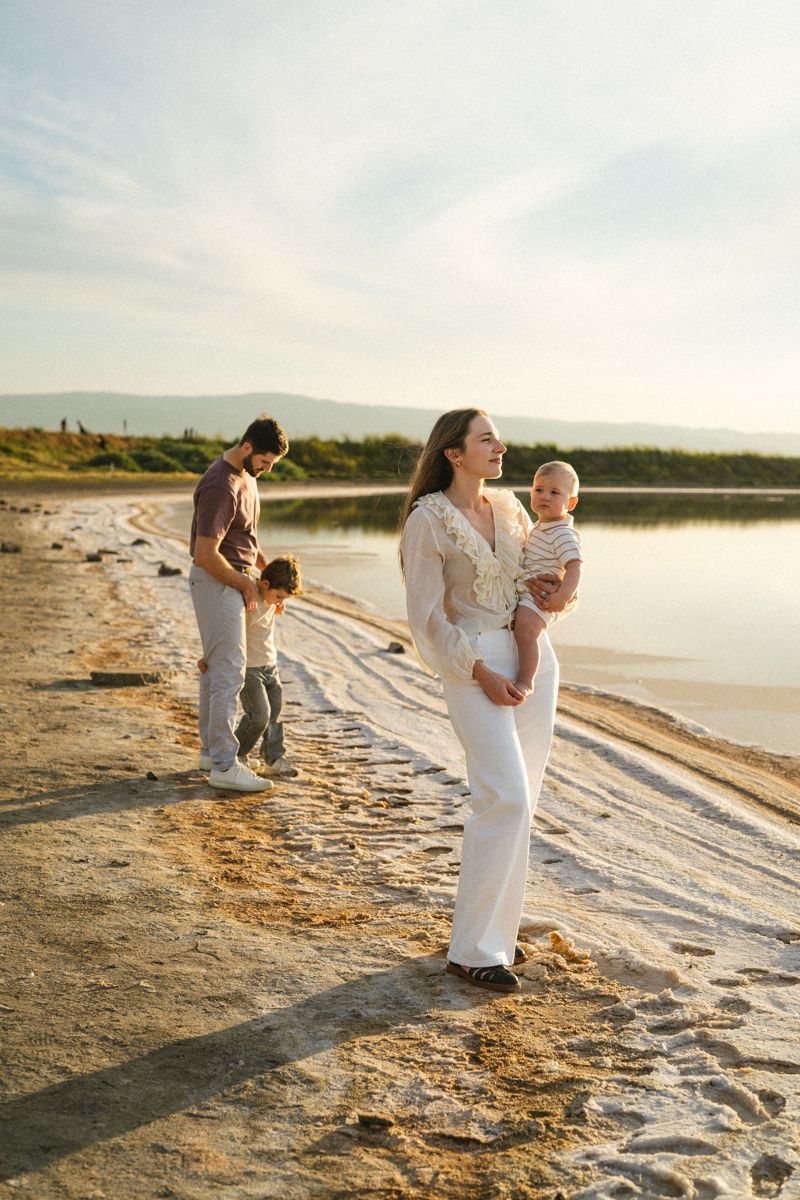 A young family enjoying a sunny day on a sandy beach by a calm lake, mother holding baby while father plays with toddler.