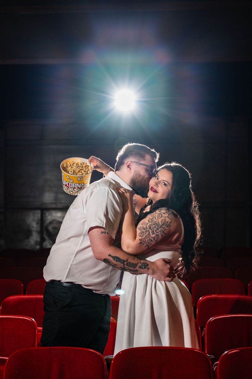 Couple embracing romantically in a movie theater with red seats, holding popcorn, backlit by a bright projector light.