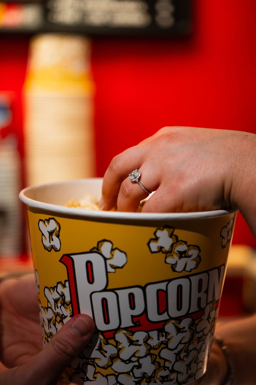 A hand with a diamond engagement ring reaching into a large yellow popcorn bucket at a movie theater.
