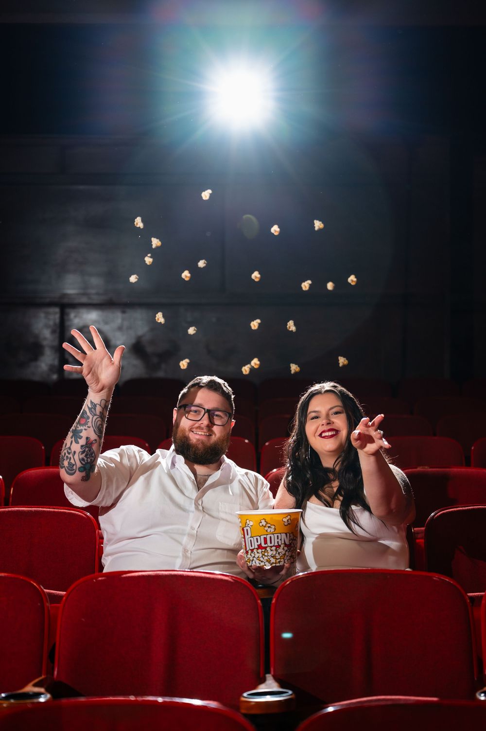 Couple laughing and tossing popcorn in red cinema seats, enjoying a fun movie date night together.