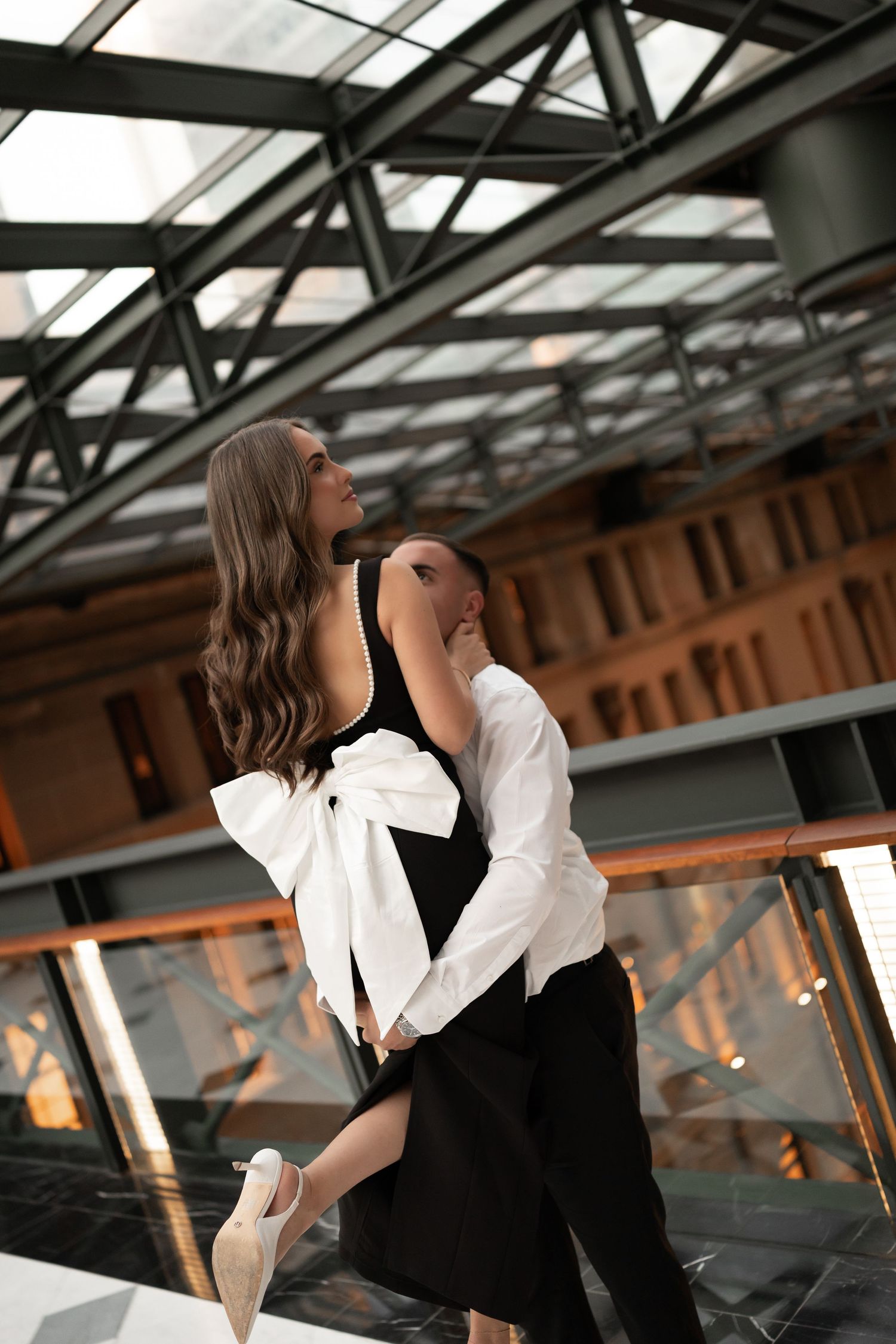 Couple embracing in elegant black and white formal attire under modern glass ceiling.