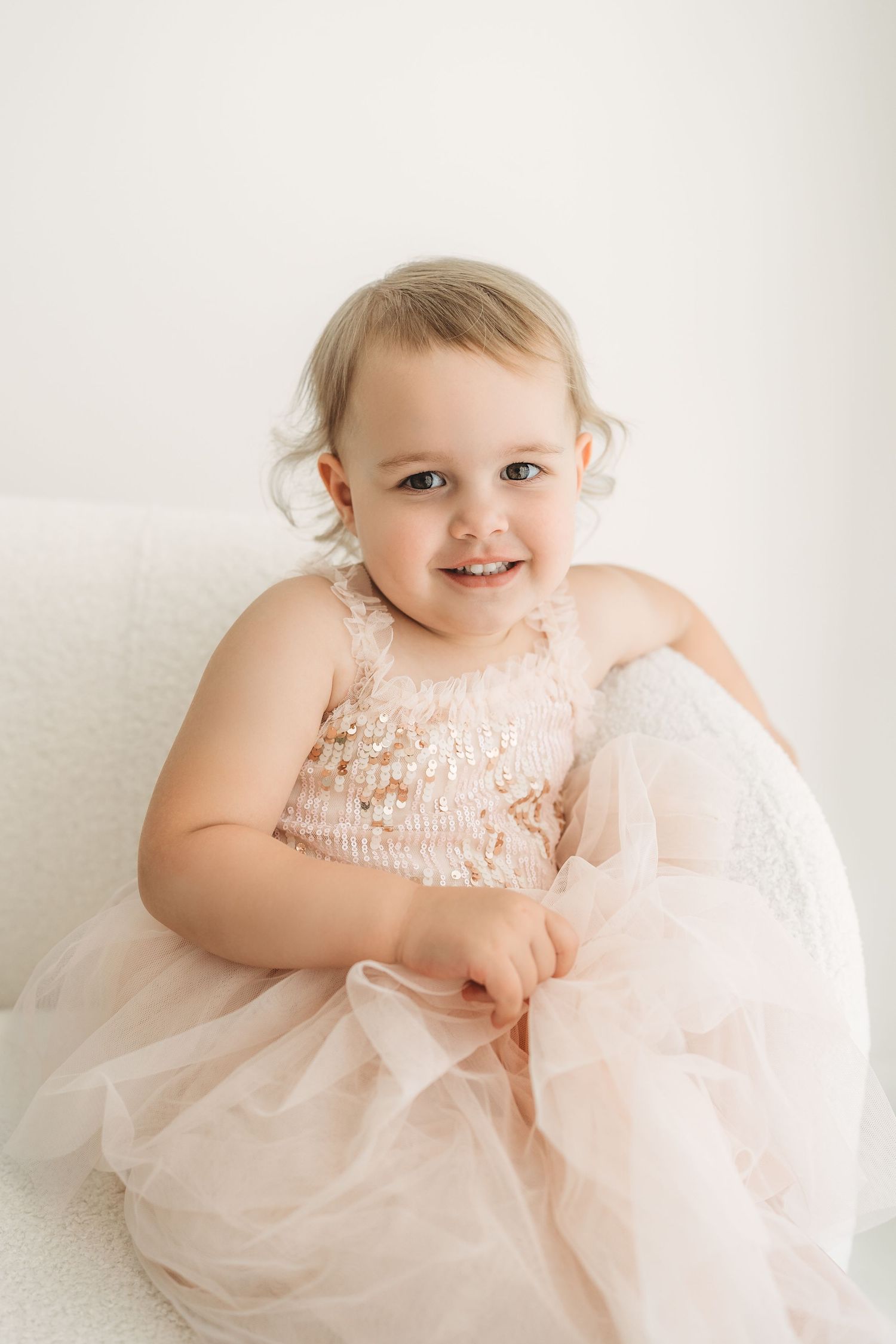 Adorable toddler girl smiling in pink sequin and tulle dress sitting on white chair.