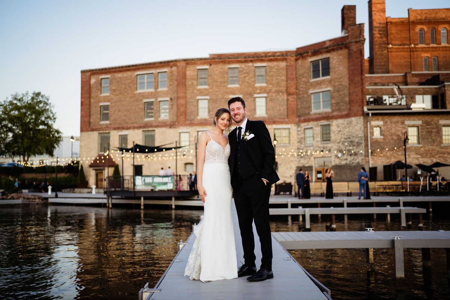 Waterfront event venue with guests gathered along a brick building dock, boats moored nearby on a sunny day. Bride and groom stand before gu