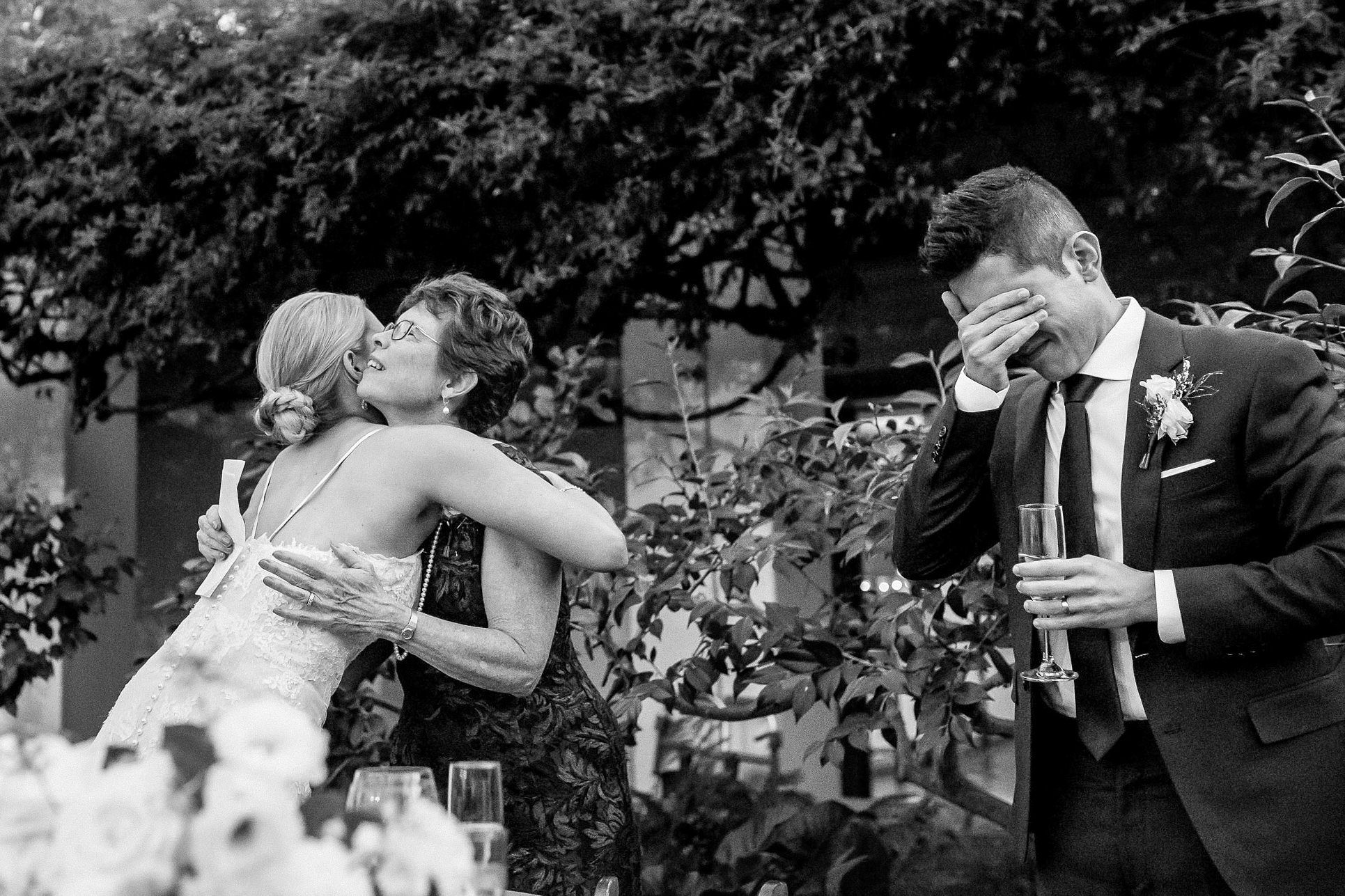 Black and white wedding photo of bride hugging a guest while emotional groom wipes tears holding champagne glass outdoors.