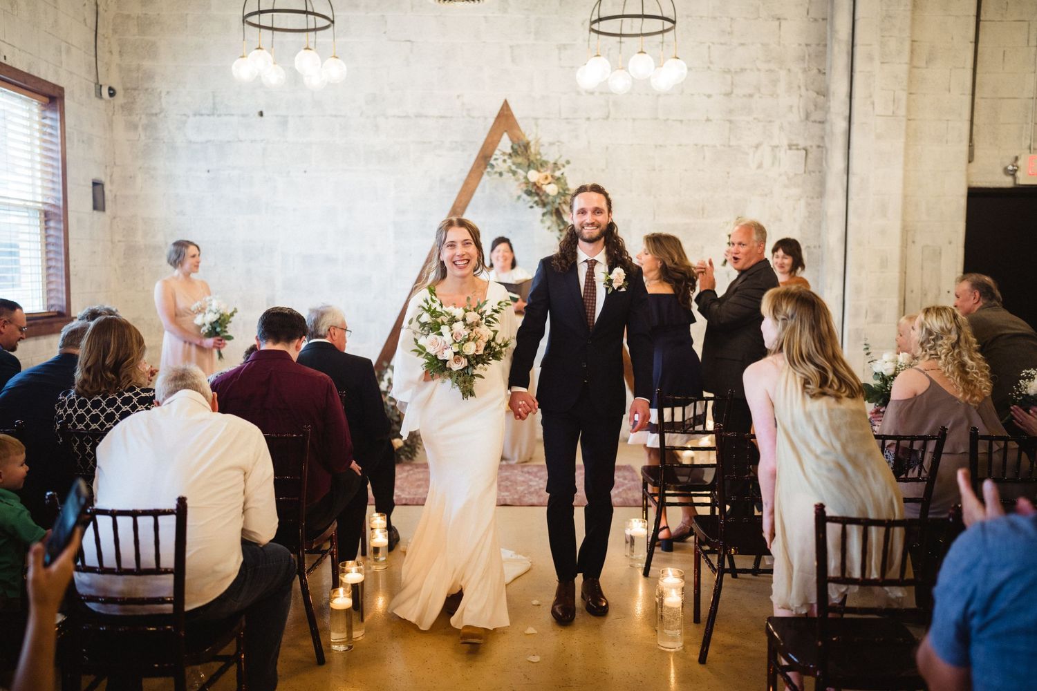 Bride and groom walk down the aisle hand-in-hand at a modern industrial wedding venue with a wooden arch.