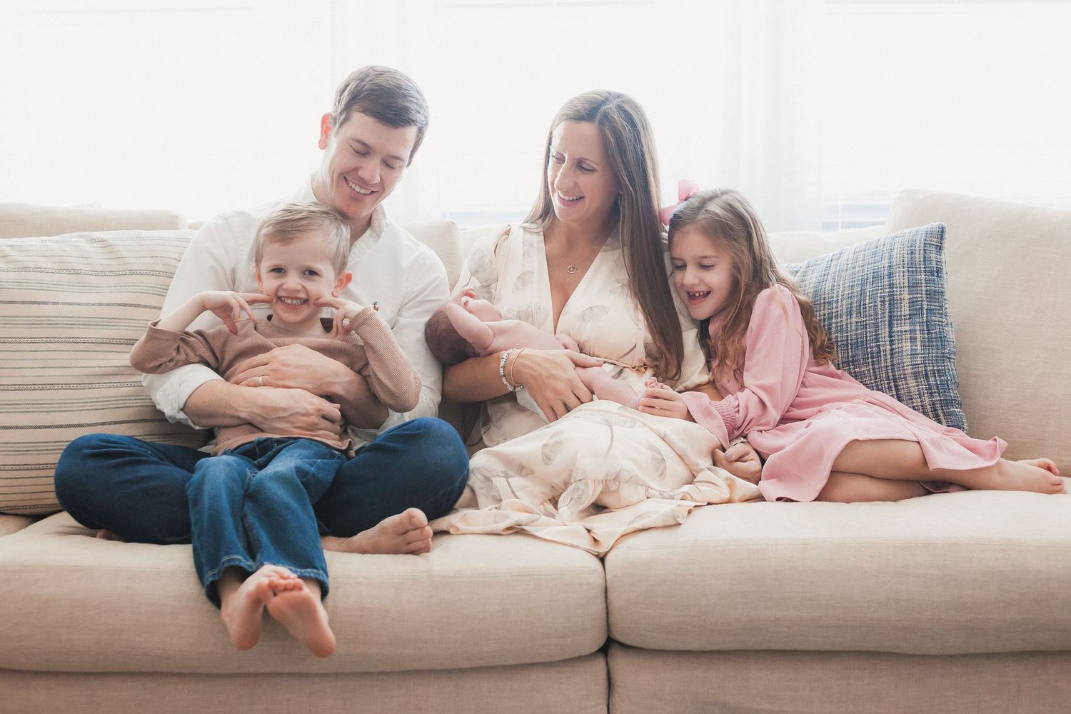 Happy family of four sitting together on a couch, parents holding a newborn baby.