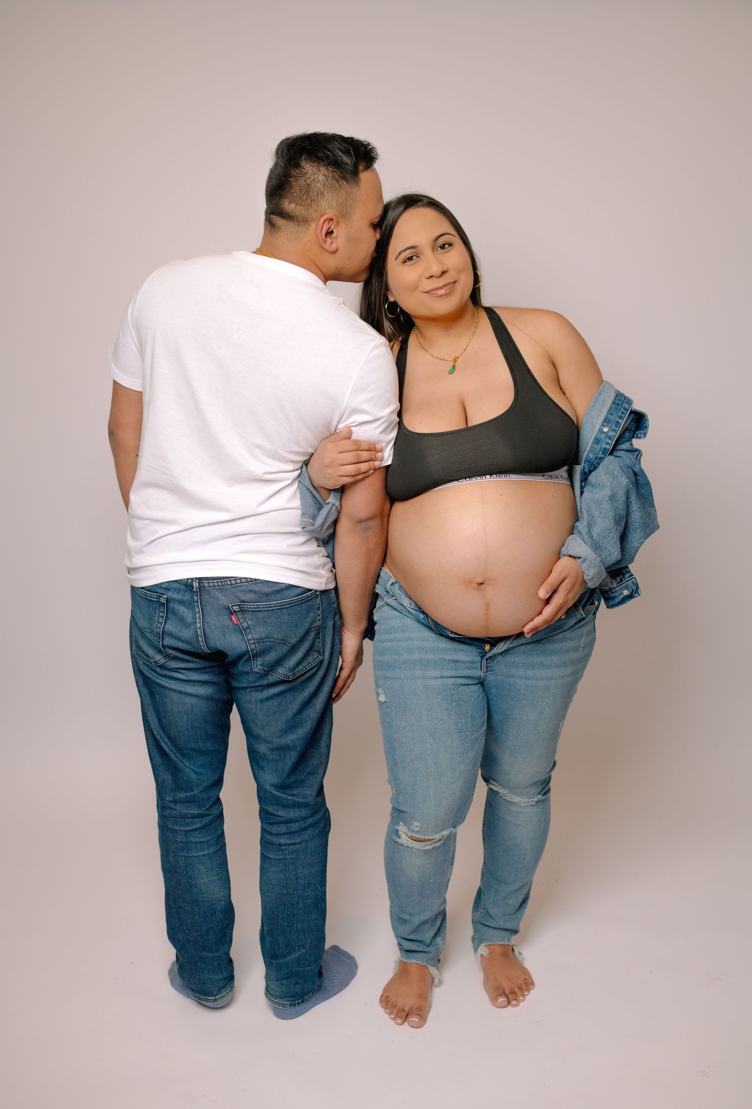 Pregnant couple posing in studio, man kissing woman's cheek while she cradles her baby bump.
