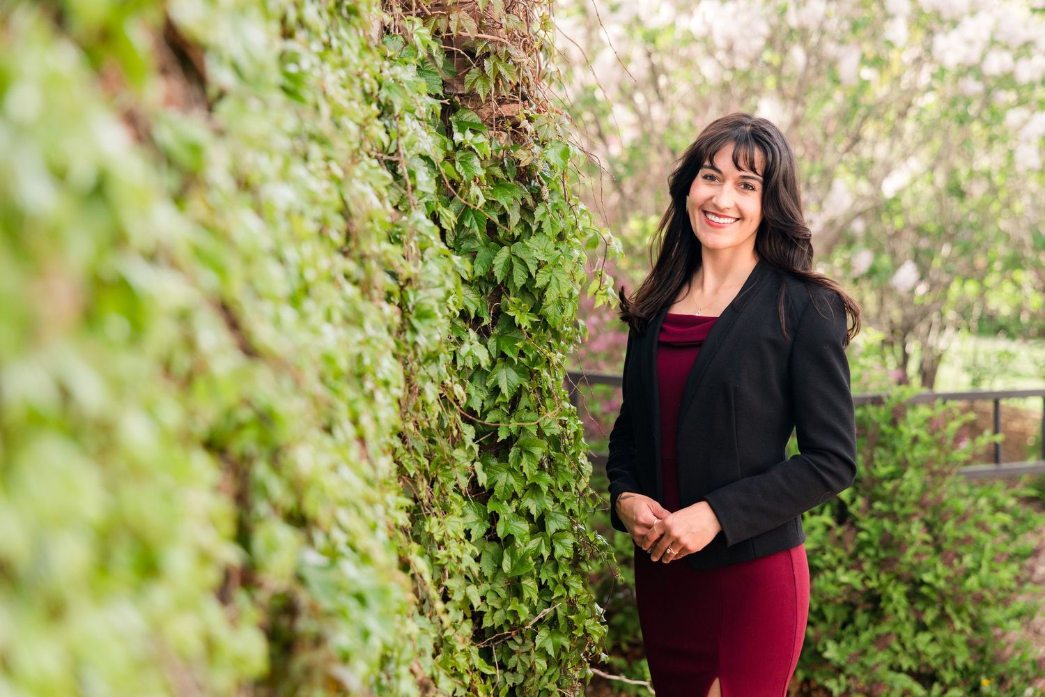 Woman in black blazer and burgundy outfit smiling beside a lush ivy-covered wall outdoors.