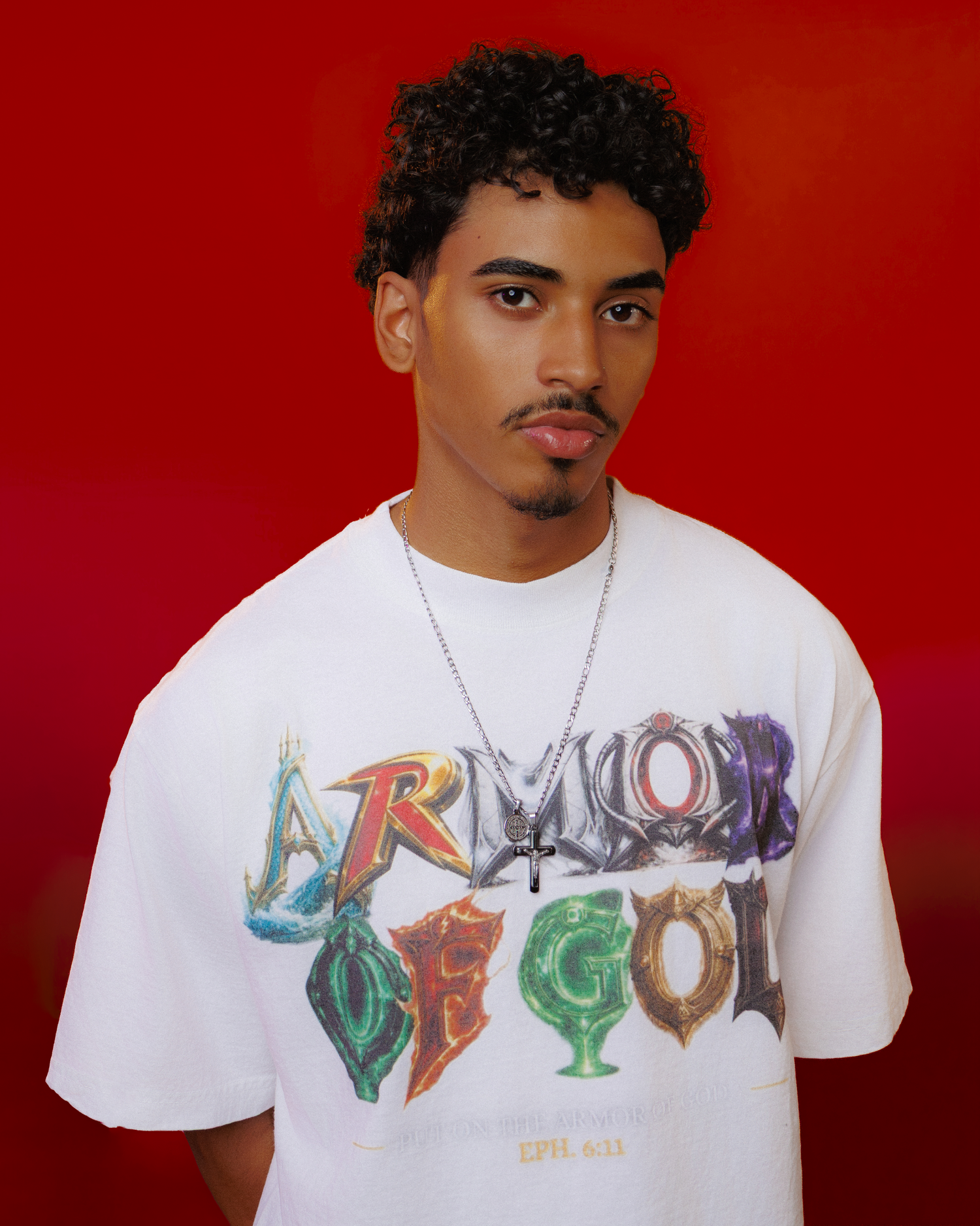 Young man wearing white Armor of God graphic tee with cross necklace against orange gradient background.