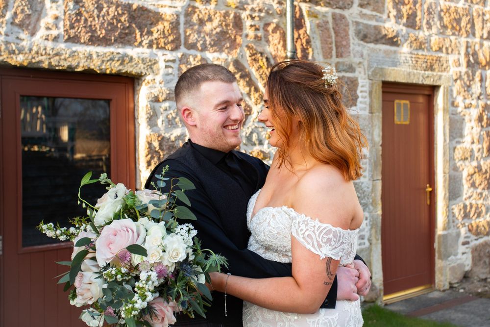 Smiling groom in black suit embraces bride in off-shoulder lace gown holding blush floral bouquet outside stone building.