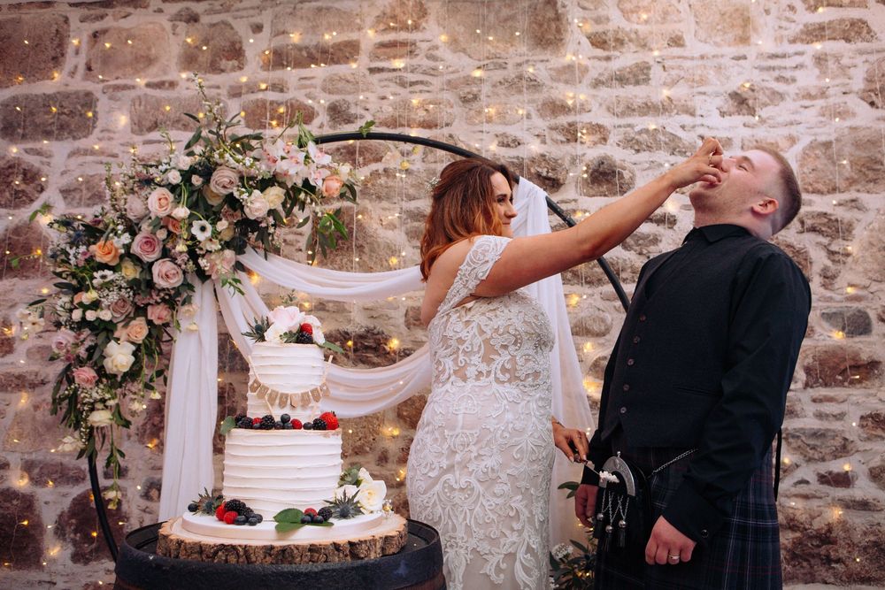 Bride feeding cake to groom in kilt at rustic stone wall wedding venue with floral hoop backdrop.