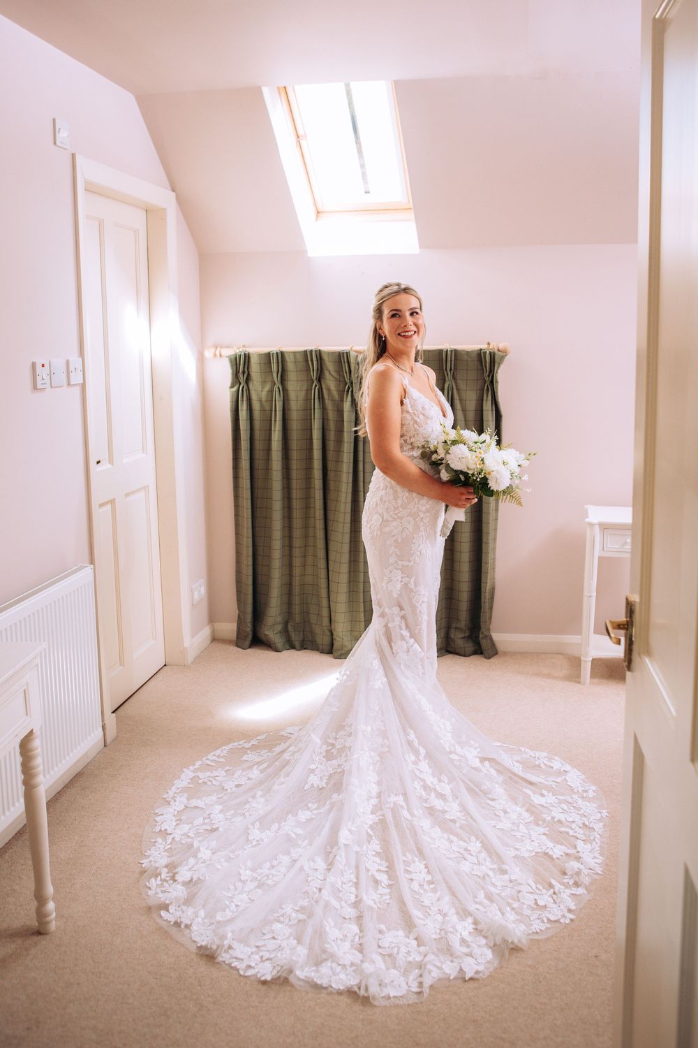 Bride in a white lace mermaid wedding gown holding a bouquet, standing in a bright room with green curtains.