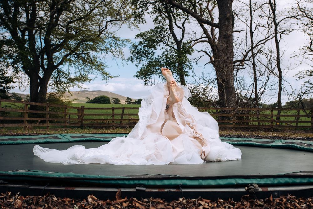 Bride in a flowing white wedding dress sitting on a trampoline outdoors surrounded by trees and green fields.
