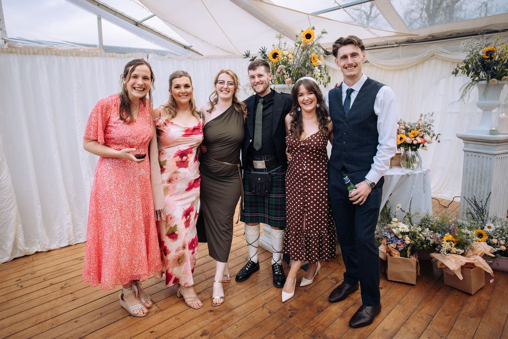 Six guests smile together in a floral-decorated marquee at a wedding, one man wearing a traditional Scottish kilt.