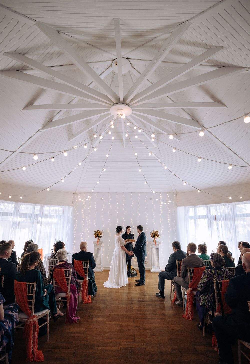 Elegant indoor wedding ceremony in a white octagonal venue with string lights, wooden floors, and guests seated in gold chairs.