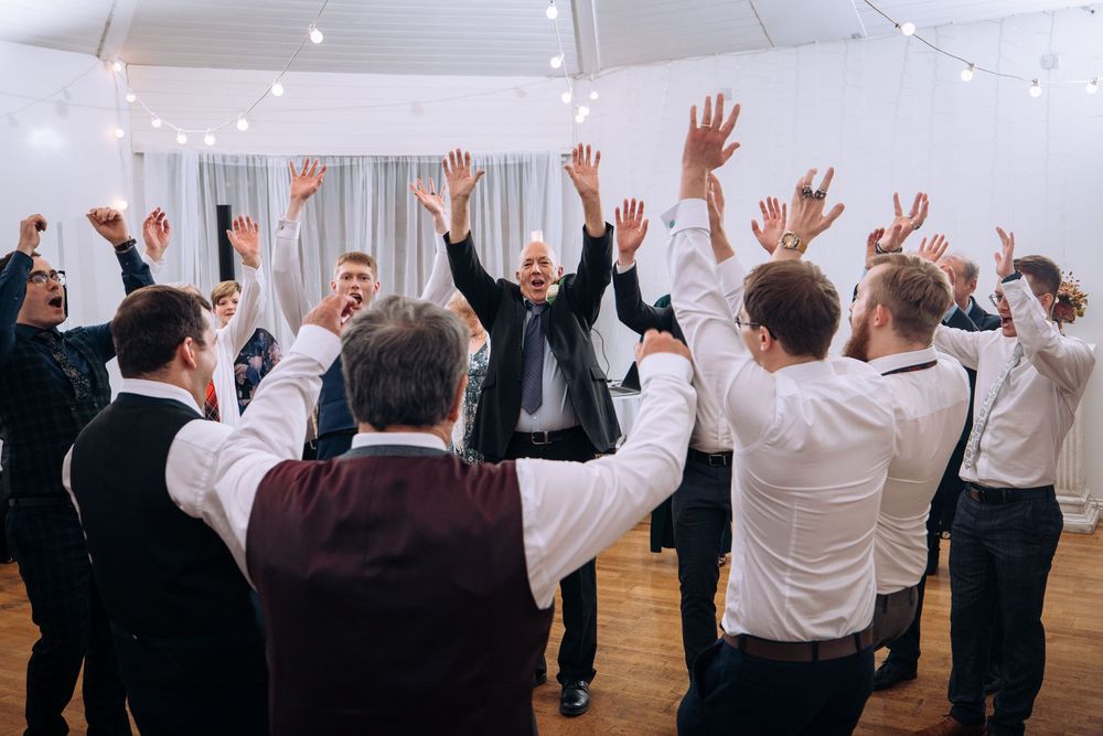 Groom celebrates with groomsmen raising hands in joyful dance at elegant wedding reception venue.
