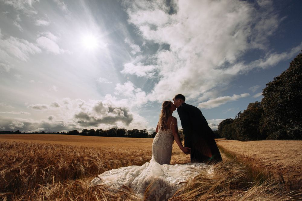 Bride and groom share a romantic kiss in a golden wheat field under a dramatic cloudy sky on their wedding day.