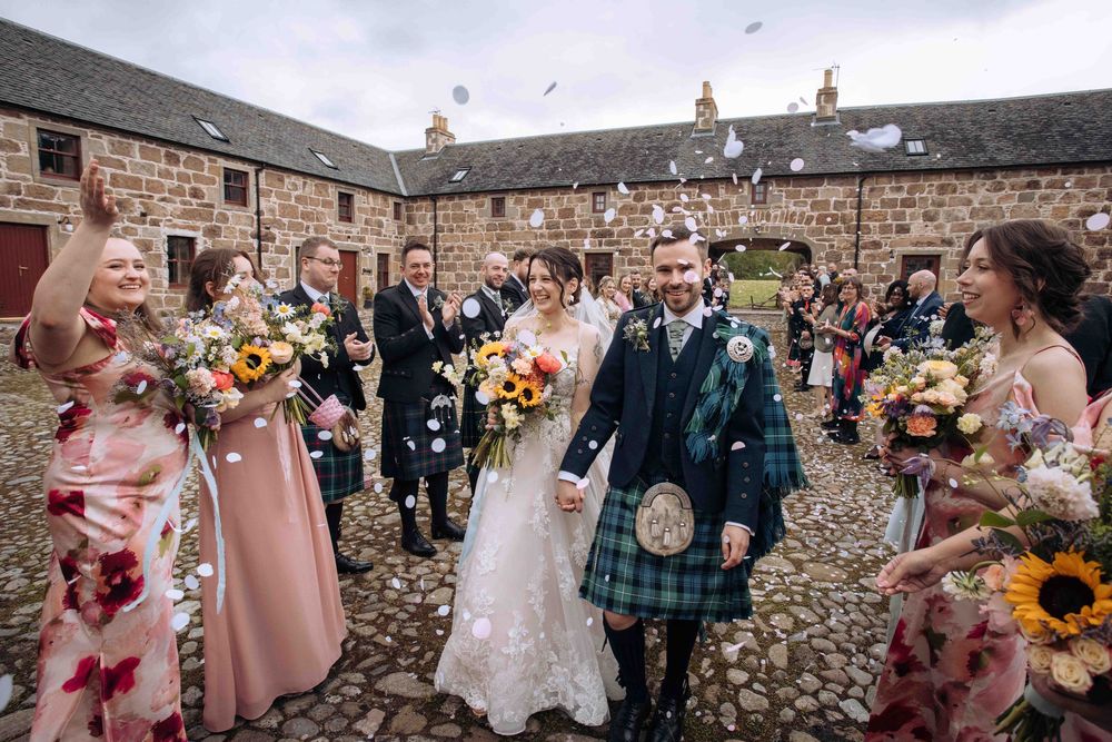 Bride and groom walk through confetti shower at Scottish wedding, groom in tartan kilt, bridesmaids in pink floral gowns.