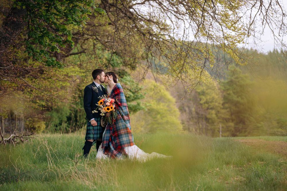 Bride and groom kissing under a tree in a lush green field, groom wearing a tartan kilt, bride holding a colorful bouquet.