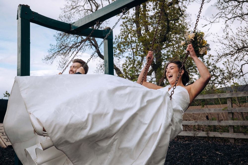 Two brides in white wedding gowns laughing joyfully while swinging on outdoor swings on their wedding day.