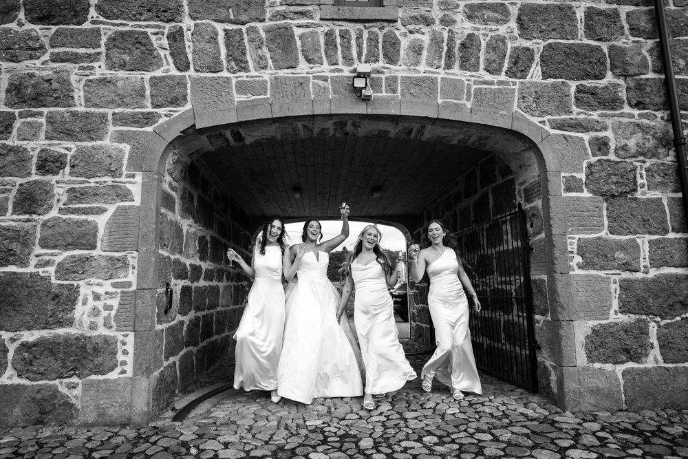 Black and white photo of four bridesmaids in white gowns celebrating under a stone archway on cobblestones.