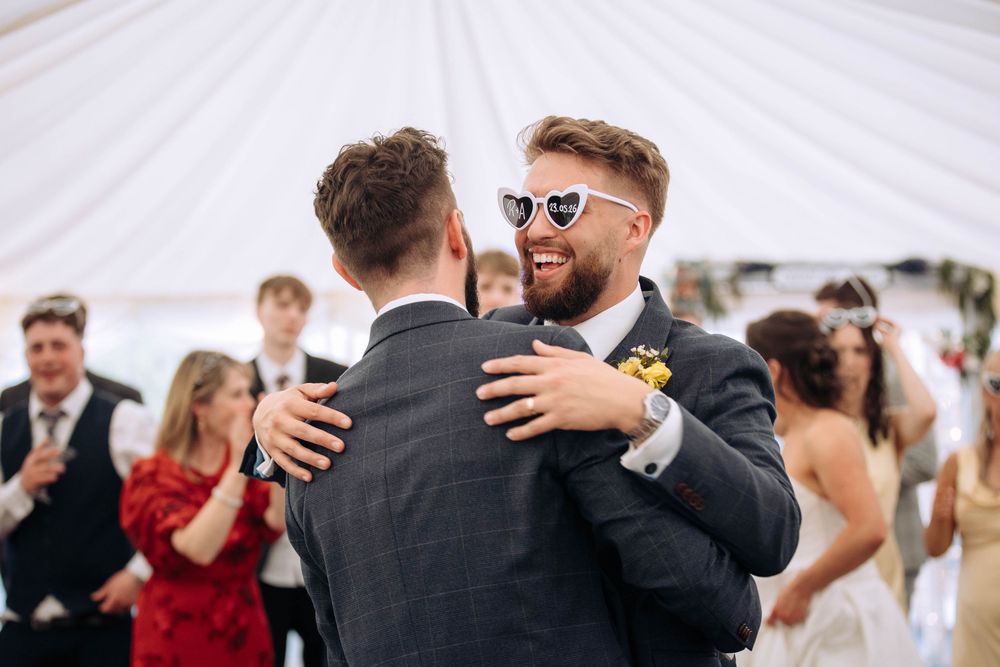Two grooms share a joyful dance at their wedding reception, one wearing heart-shaped sunglasses and a boutonniere.