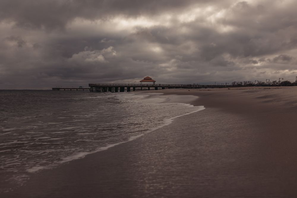 Moody beach pier with red pavilion under dramatic cloudy sky, calm waves on sandy shore at dusk.
