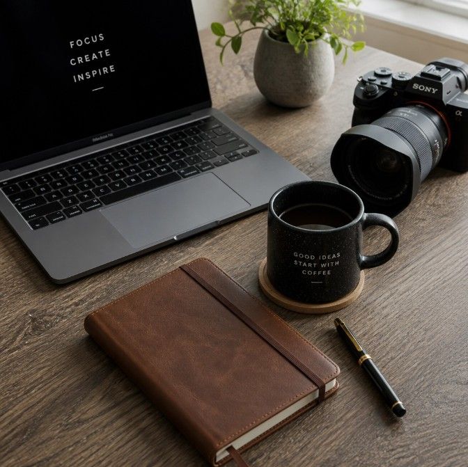 A flat lay workspace with a laptop, Sony camera, leather journal, pen, and coffee mug on a wooden desk.