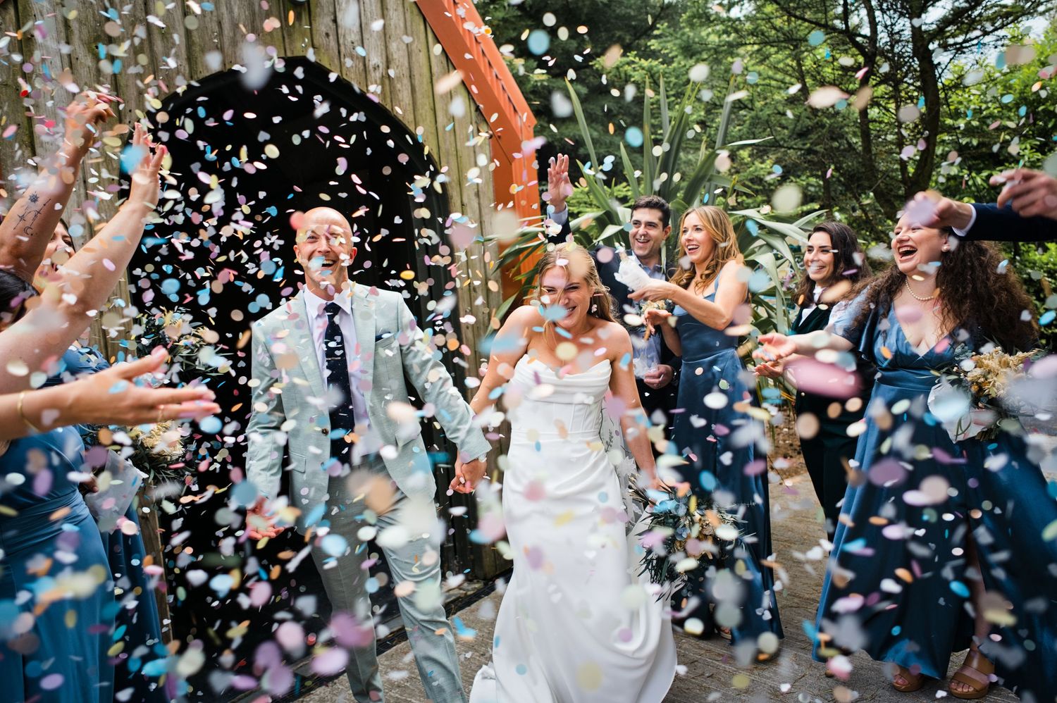 Joyful newlyweds walk through colorful confetti shower surrounded by smiling guests in blue attire at outdoor wedding celebration.