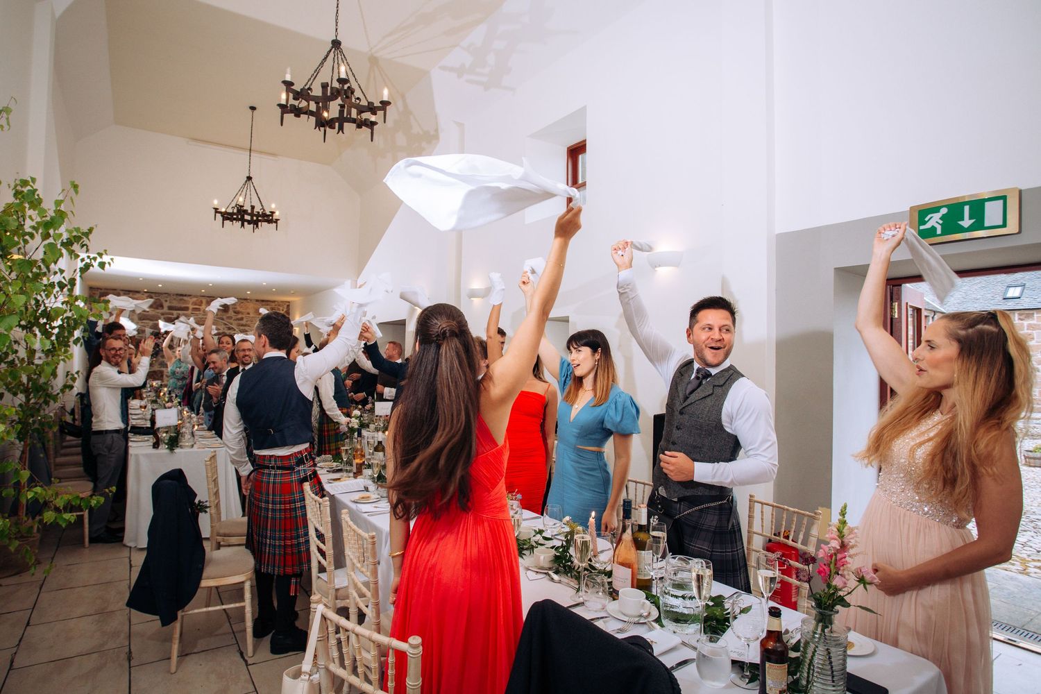 Wedding guests celebrate joyfully waving napkins at an elegant reception venue with floral table decorations.