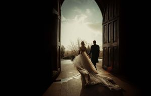 Bride and groom silhouetted in grand church doorway, wedding gown trailing elegantly as they gaze at scenic outdoor landscape.