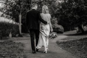 Black and white photo of bride and groom walking arm in arm down a path, bride holding a bouquet.