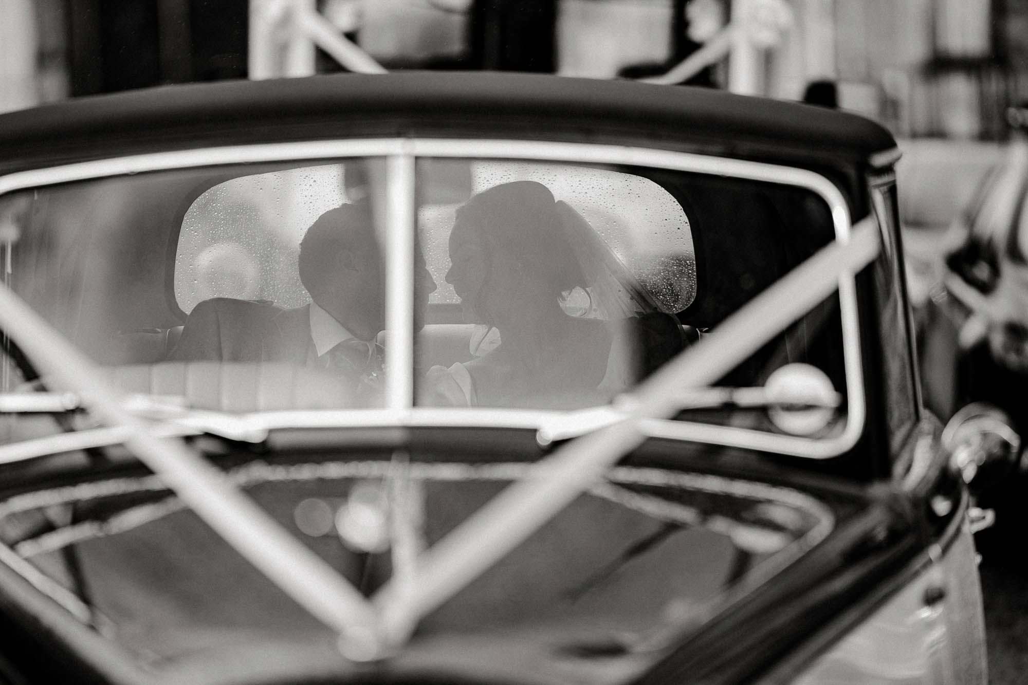 Black and white photo of a couple sharing a kiss inside a vintage classic car, captured through the windshield.